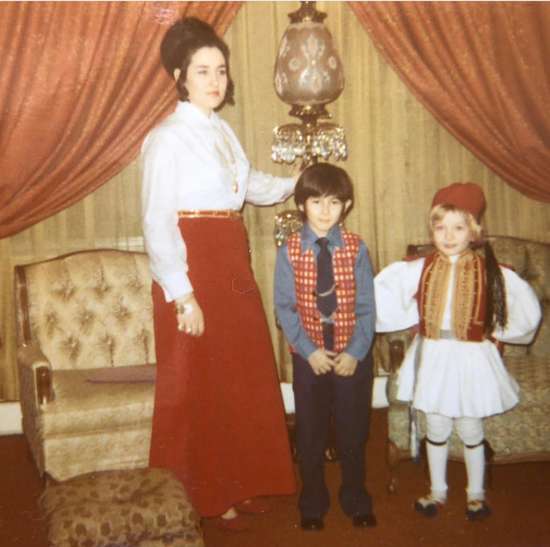 Vintage childhood photograph of Gregory Pappas on the right wearing an evzone costume, standing beside two other people in a living room.