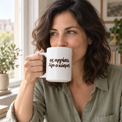 AI-generated lifestyle image: woman drinking coffee in a sunlit kitchen with the Ας Αργήσει Λίγο ο Κόσμος white 15 oz mug